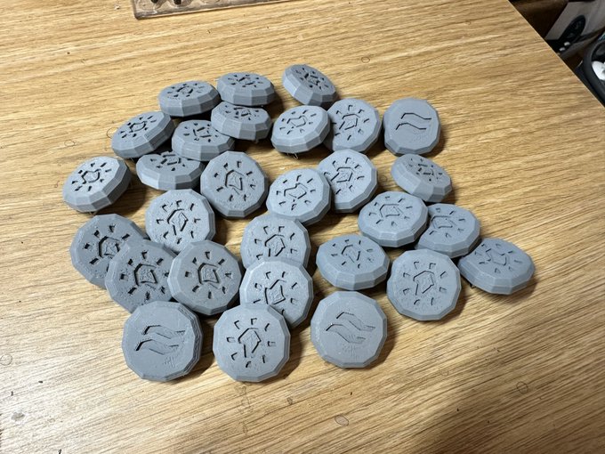 A collection of approximately 25 gray 3D-printed tokens with various symbols and rune-like designs scattered on a wooden table surface.