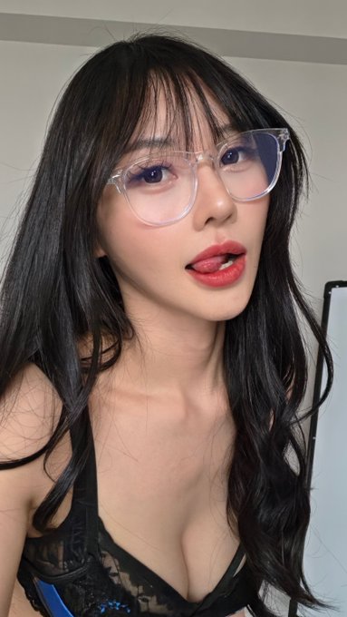 A young woman with long dark hair and bangs wearing clear-framed glasses, red lipstick, and a black lacy top, photographed indoors.