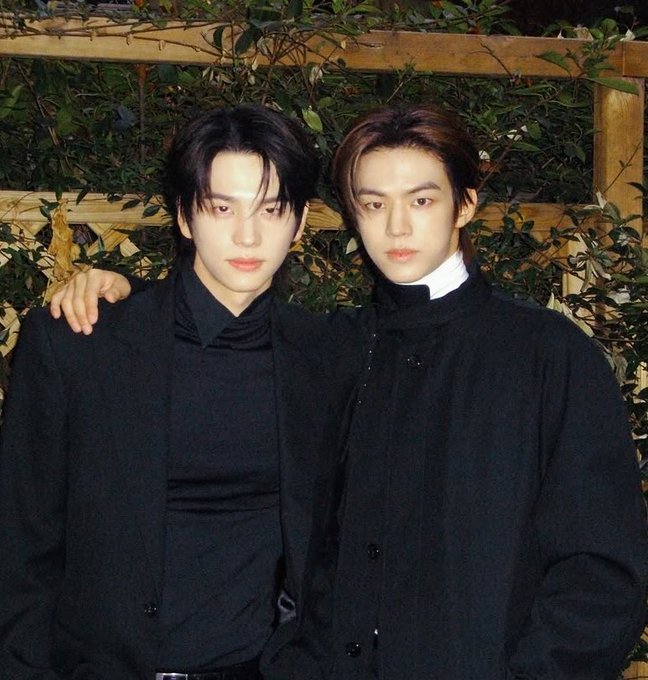 Two young men in all-black outfits stand close together, with one resting his arm on the other's shoulder in front of a foliage-covered background.