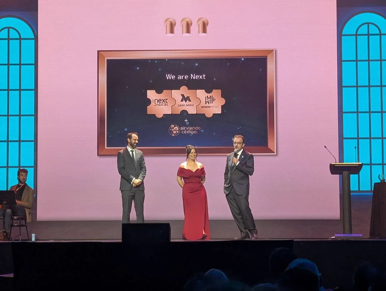 Fotografía del escenario de la gala Sirviendo Código con los presentadores muy elegantes: Isabel Rodríguez luciendo un vestido largo rojo, acompañada por Pablo López y Ramsés Rodríguez en esmoquin y corbata. Al fondo se observa la pantalla corporativa con el mensaje "We are Next" junto a los logotipos de los colaboradores.