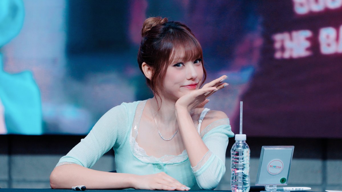 A young woman with her hair in a bun sits at a table, smiling gently while resting her chin on her hand, with a water bottle and a nameplate visible on the table.