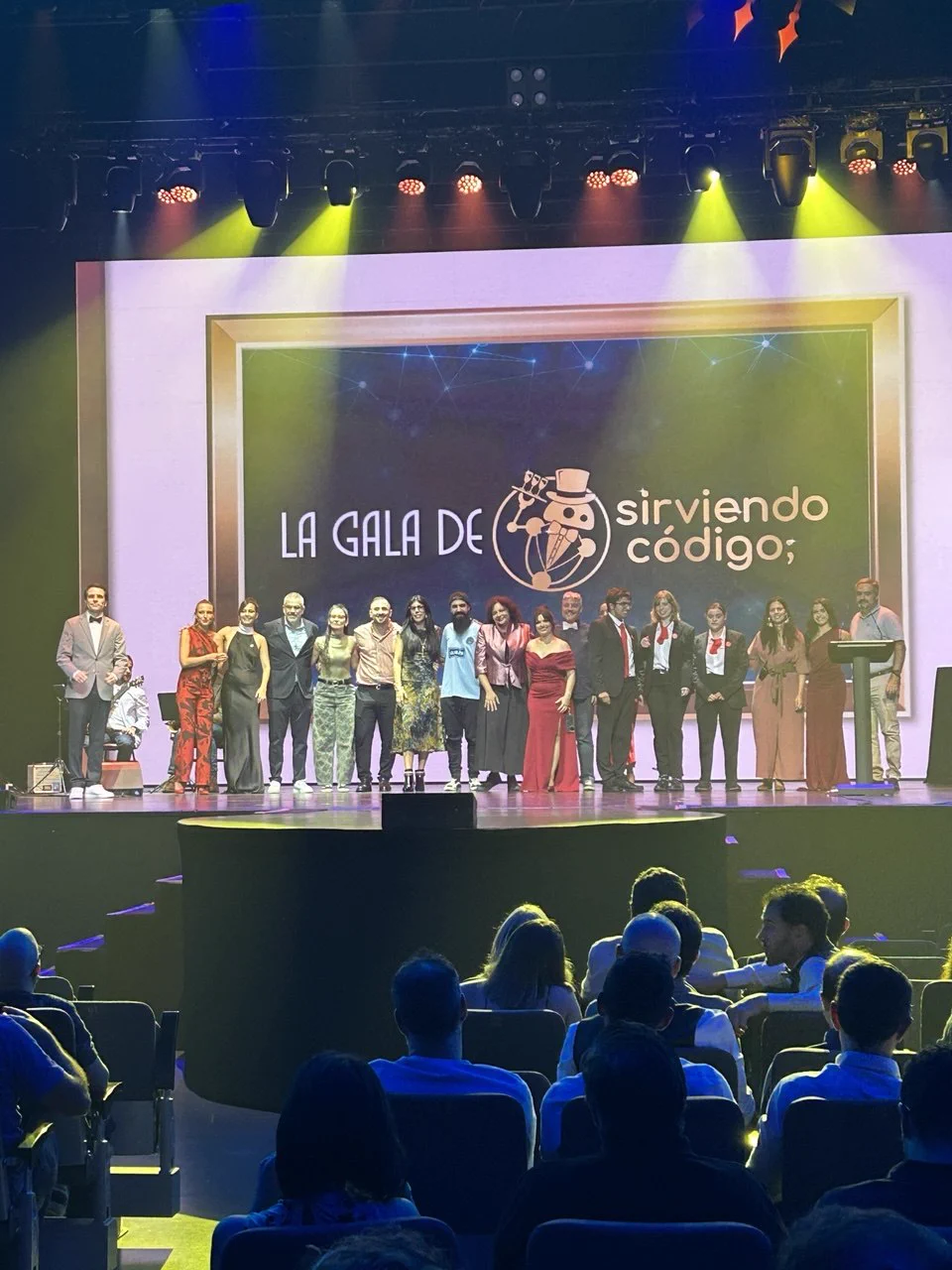 Fotografía panorámica del escenario iluminado de la gala Sirviendo Código. Todo el equipo organizador posa alineado, muy bien vestido y con grandes sonrisas de agradecimiento hacia el público. De fondo destaca la pantalla principal con la gráfica del evento "La Gala de Sirviendo Código".