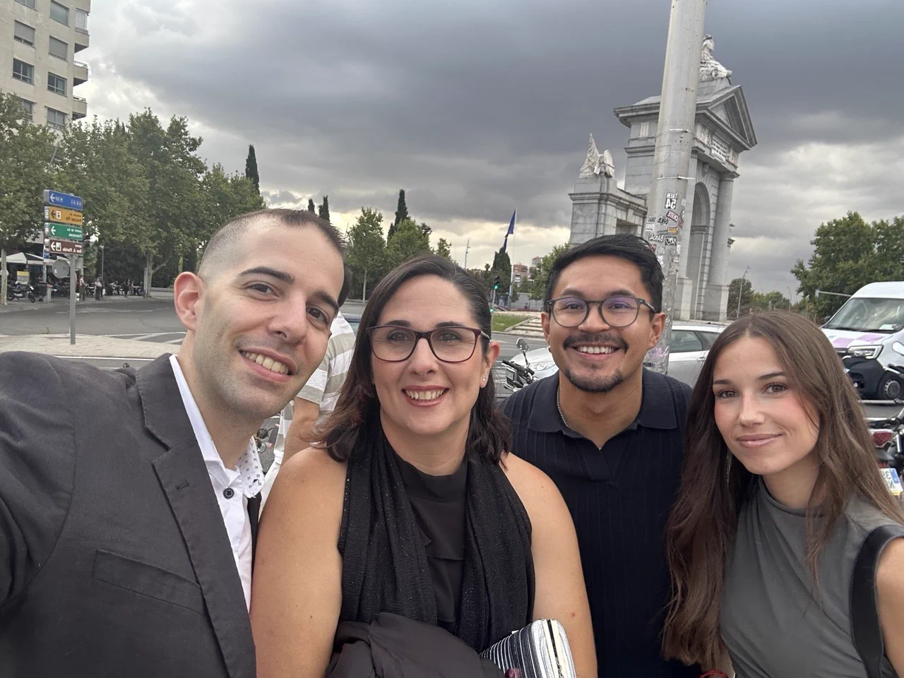 Fotografía tipo selfie de cuatro compañeros de Lean Mind posando sonrientes y vestidos para la gala en el exterior, antes de entrar. En primer plano y de izquierda a derecha, se encuentran Adrián Ferrer, María Blé, Mario Pinto y Aymara Sánchez. Al fondo se distingue la Puerta de Toledo en Madrid bajo un cielo nublado.