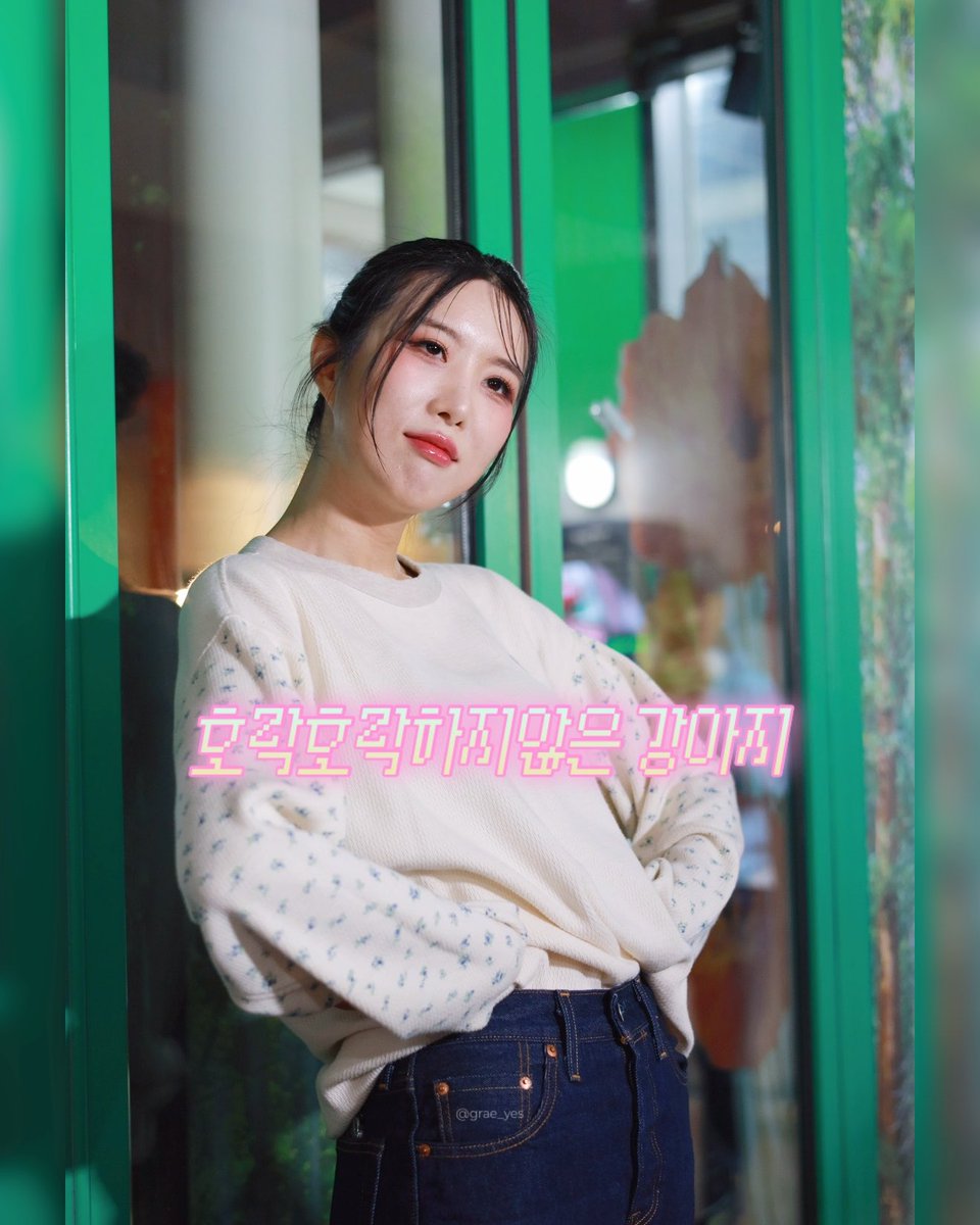 A portrait of a young woman with dark hair wearing a cream-colored sweater and blue jeans, leaning against a bright green door frame, with Korean text '흔들흔들가지 않는 강마지' overlaid in pink and white.