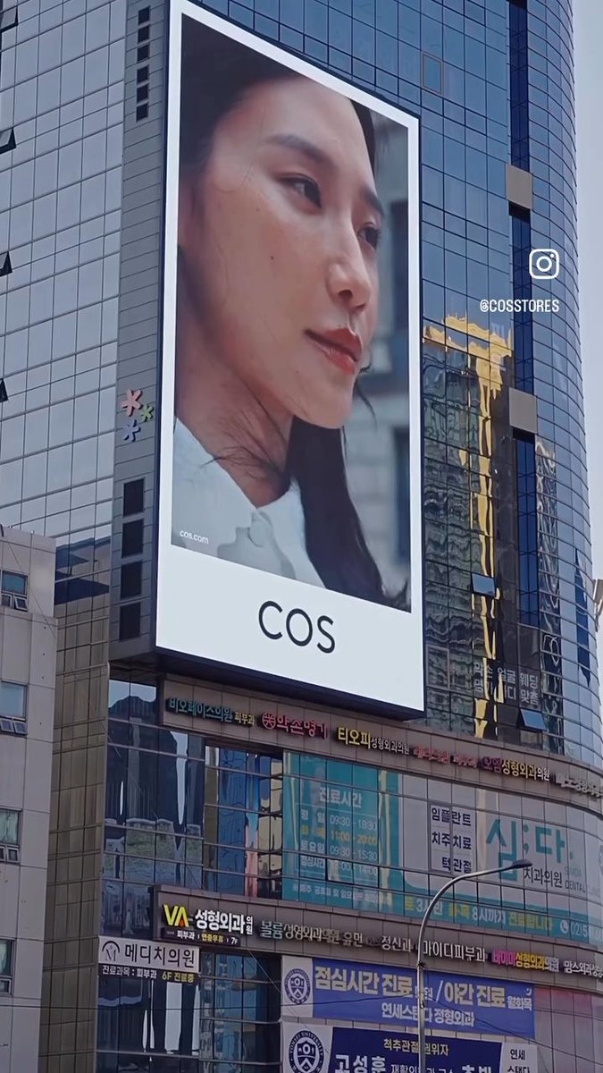 A large outdoor billboard advertising 'COS' displays a close-up portrait of a woman's face, mounted on a glass building with Korean signage below, tagged @cosstores on Instagram.