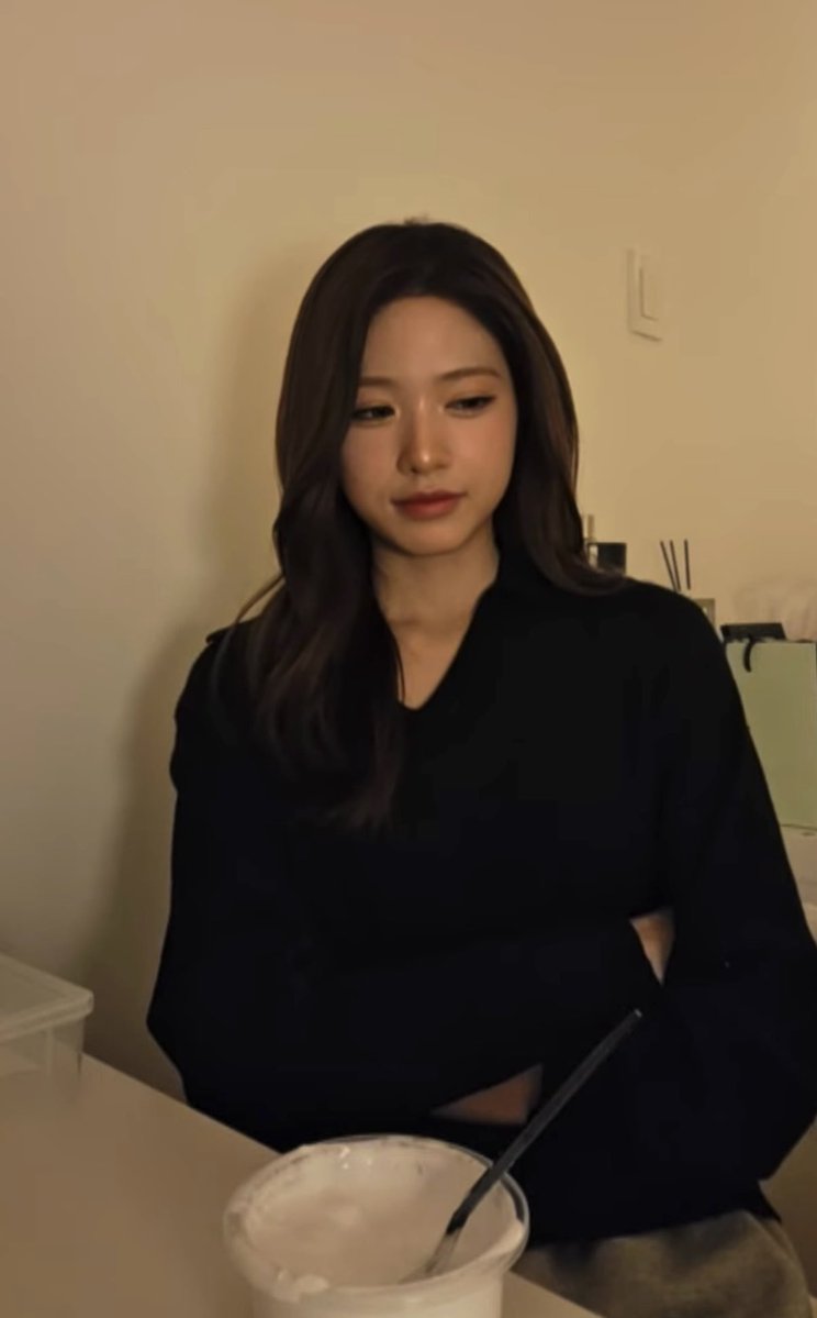 A woman with long dark hair wearing a black top sits at a table with a plastic cup containing a drink and straw in front of her.