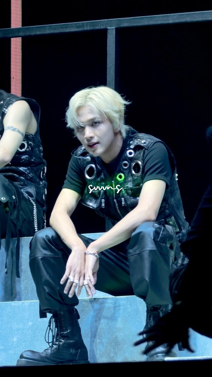 Concert photo of a young male performer with blonde hair wearing a black leather vest with metal ring details, black pants, and combat boots, seated on stage steps with his hands resting on his knees.