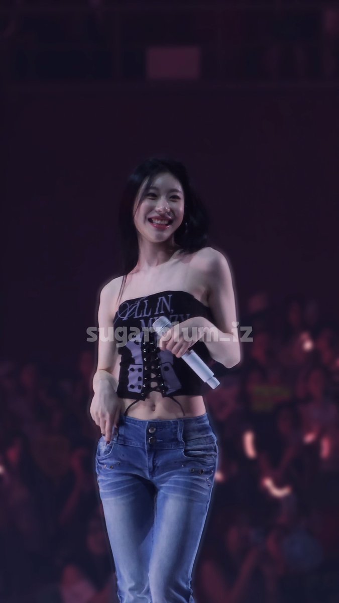 A woman with dark hair smiles on stage while holding a microphone, wearing a black corset-style top with 'ALL IN' visible and blue jeans, with a watermark reading 'sugarplum_iz' across the image.