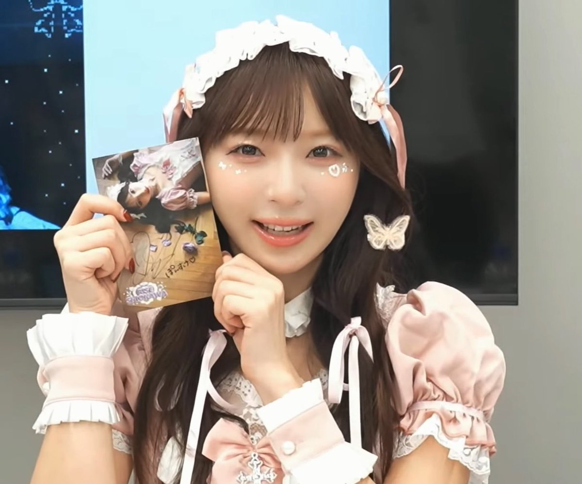 A smiling young woman in a pink and white maid-style outfit holds up a signed photo card, wearing cute face decorations and a butterfly hair accessory.