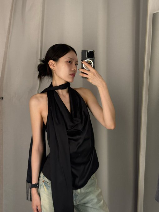 A mirror selfie of a woman wearing a black halter-style top with a long flowing tie around her neck, paired with light blue jeans.