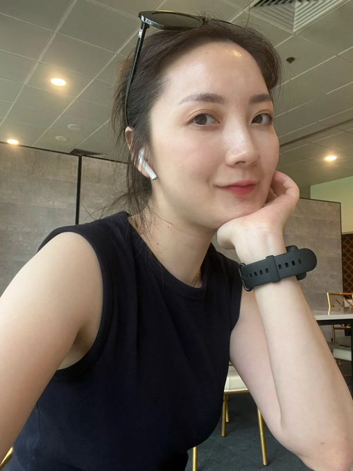 A young woman with sunglasses perched on her head and wireless earbuds in poses for the camera with her chin resting on her hand, wearing a black sleeveless top and a smartwatch, in what appears to be a casual cafe or restaurant setting. The image captures a relaxed, confident selfie moment that reflects the bold, carefree spirit described in the post about feeling emboldened to share freely on X.