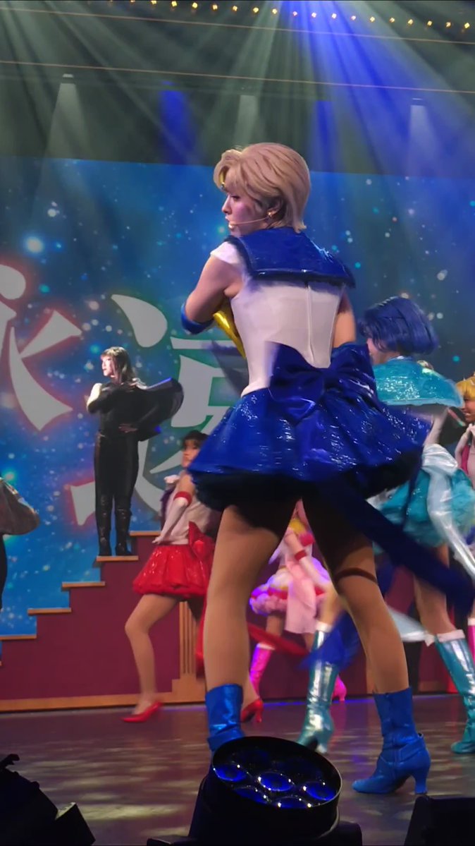 A stage performer in a Sailor Moon-inspired blue and white sailor costume and blonde wig stands on stage, with other performers in various costumes visible in the background under theatrical blue lighting.