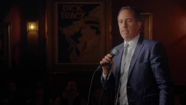 A man in a suit performs stand-up comedy on stage, holding a microphone in front of a "Dick Tracy" poster, with audience members visible behind him. The dimly lit club setting and the performer's mid-sentence expression suggest he's delivering a punchline about "millions," tying into the caption about large sums of money.