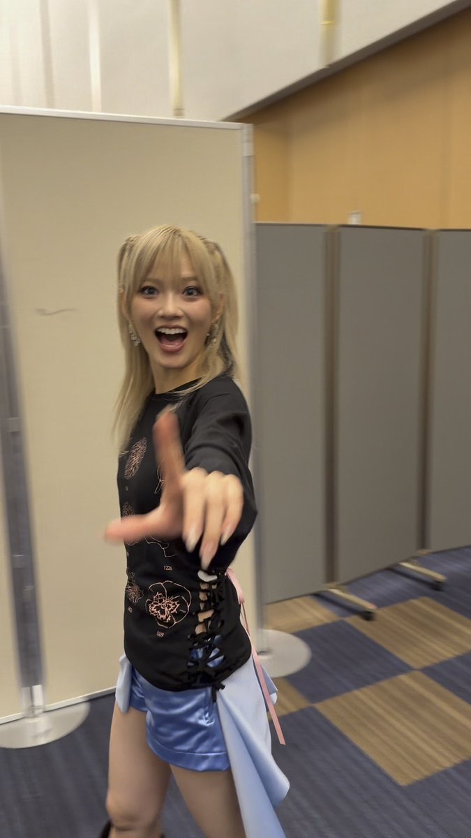 A young woman with blonde hair, wearing a black corset-style top and light blue skirt, playfully reaches toward the camera with an excited expression in what appears to be a backstage area, with the caption playfully calling out her funny face and tagging #鶴屋美咲 (Tsuruya Misaki).