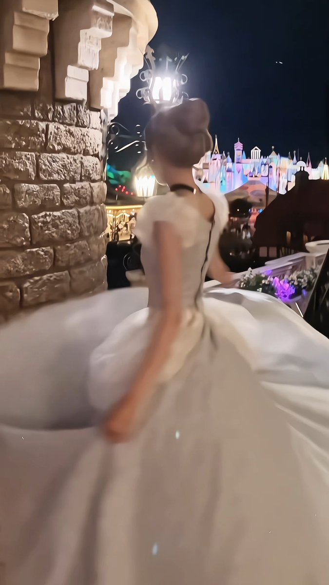 A blurred figure in a voluminous white gown spinning at night in front of an illuminated castle, with a stone structure and a lit lamppost nearby.