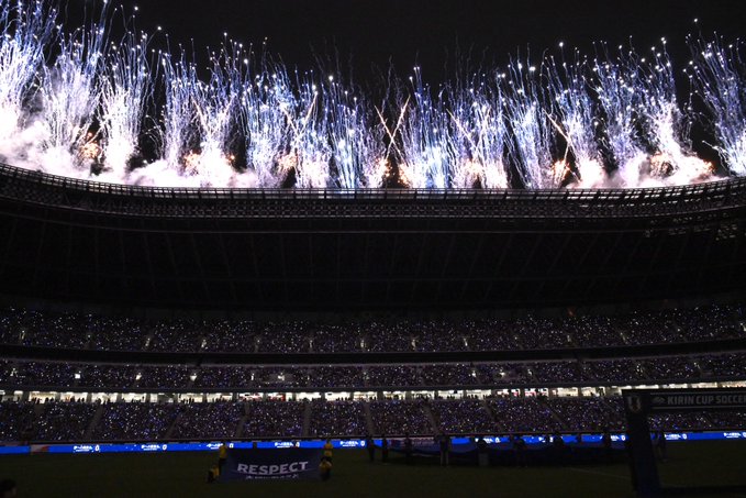 Spectacular pre-match fireworks in Japan's national team blue and white illuminate the night sky above the stadium, where a packed crowd of supporters shines their lights during the event, with "RESPECT" displayed on the pitch and a "KIRIN CUP SOCCER" banner visible on the sidelines, perfectly capturing the emotional tribute described in the caption.