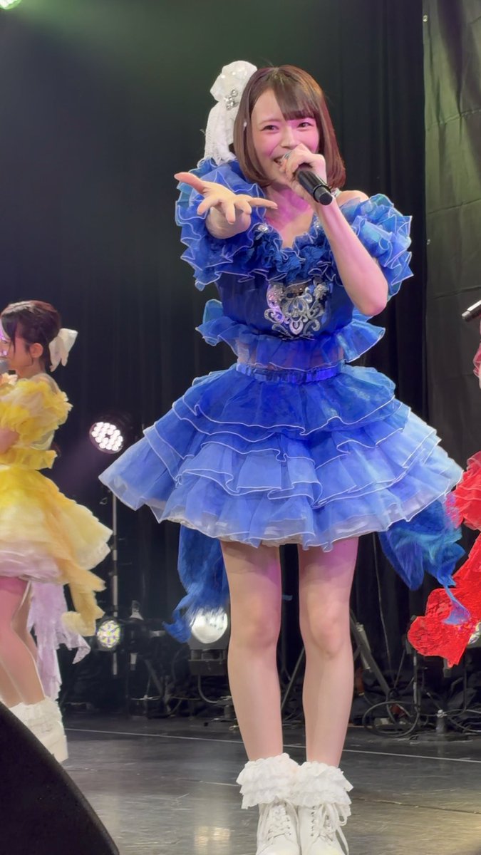 A performer with short hair wearing an elaborate blue ruffled stage dress and white boots, singing into a microphone on stage.