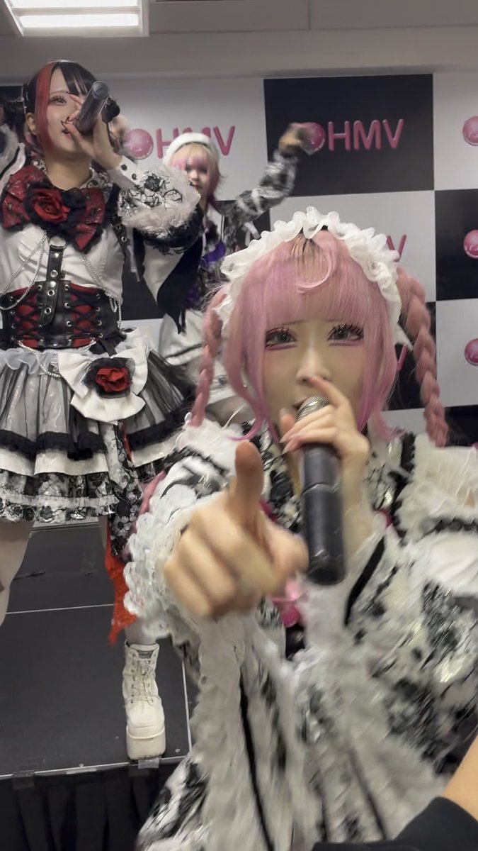 Japanese performers with pink hair are on stage in elaborate black-and-white frilly costumes; one is singing into a microphone while pointing at the viewer, and the backdrop features HMV branding.