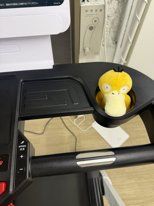 A small Psyduck plush toy sits in a cup holder on what appears to be a treadmill or exercise machine control panel, with Japanese text on a sign in the background.