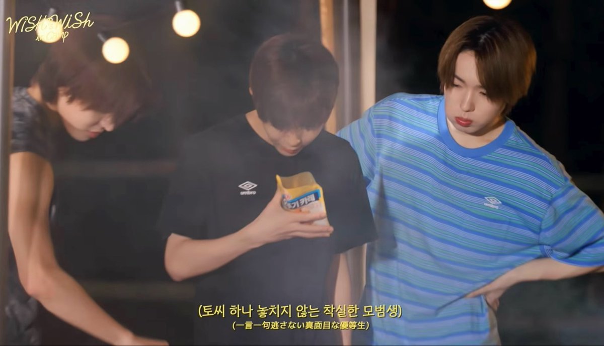 Three young men are featured in what appears to be a behind-the-scenes clip; the center figure in a black Umbro shirt examines a product, while the man on the right in a blue striped shirt looks at him; Korean subtitles read '(토씨 하나 놓치지 않는 착실한 모범생) (一言一句逃さない真面目な優等生)' with a 'WishUpon' watermark.