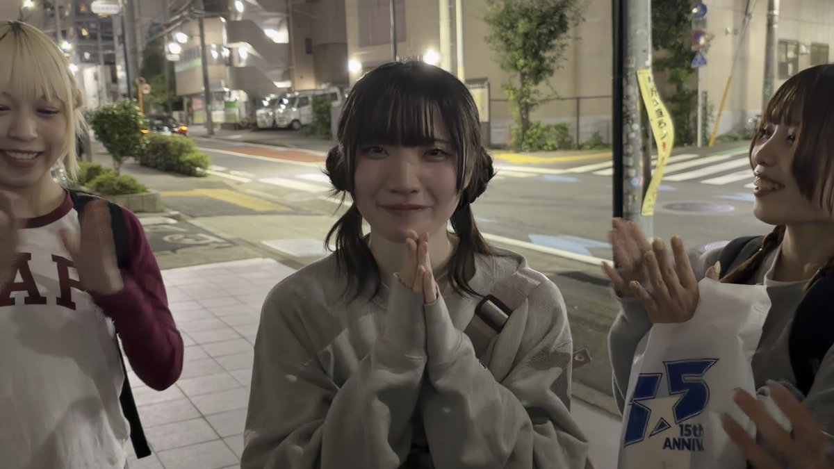 Three young women stand on a nighttime street; the central figure smiles with hands pressed together in front of her chest, wearing a light gray sweatshirt with star patterns.