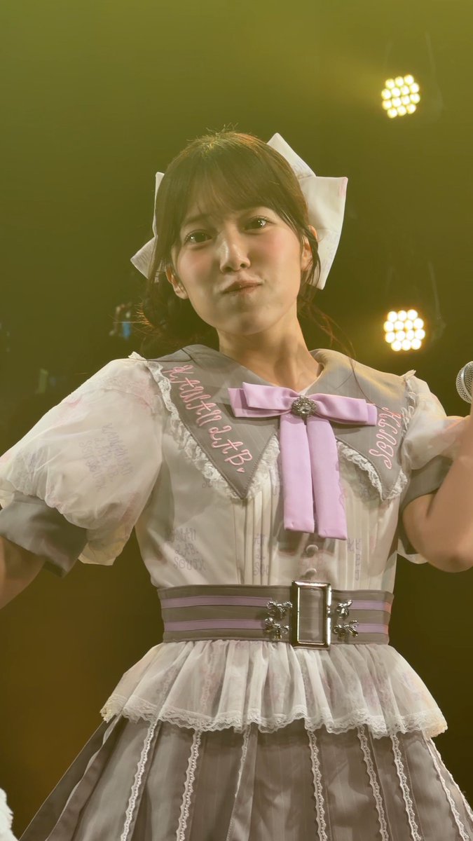 A performer on stage wears a maid-style dress with a purple bow and white headbow, with handwritten text on the collar reading "'CHEAT MASTER'" and "3LA13S".