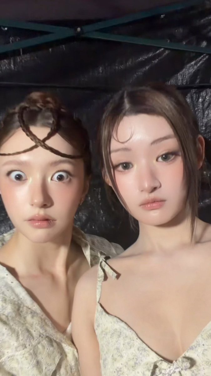 Two young women with styled hair and makeup pose closely together for a selfie in a dimly lit room; one makes a wide-eyed expression while the other has a calm, composed look.