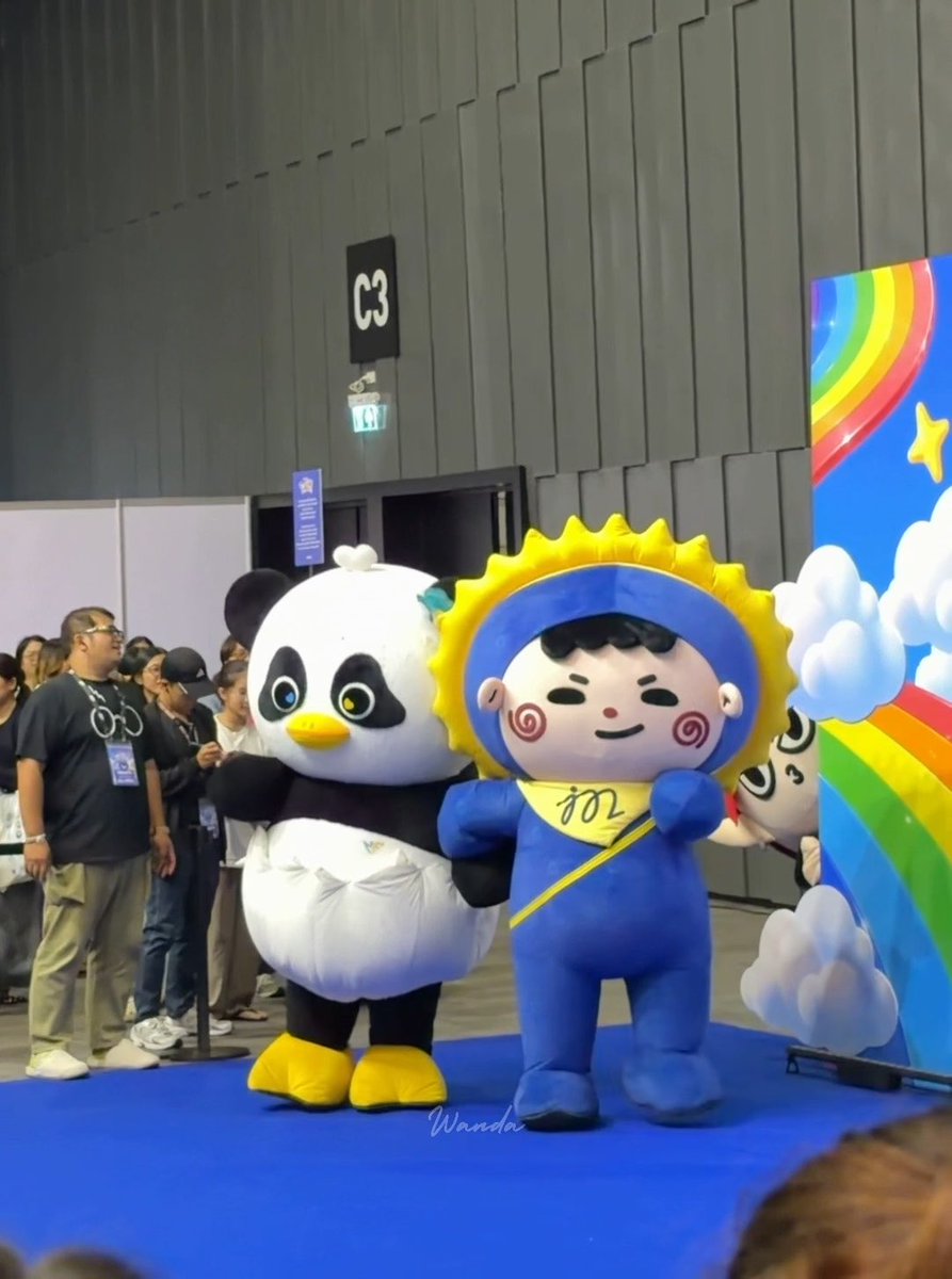 Two mascot characters, a panda and a blue figure with a yellow sunflower hood labeled "402," pose on a blue carpet in front of a sign marked "C3," with a rainbow-striped photo backdrop nearby. The playful mascots appear to be at an event, matching the post's humorous story about "Jum-Maw" and "Luna" interacting during a photo session.