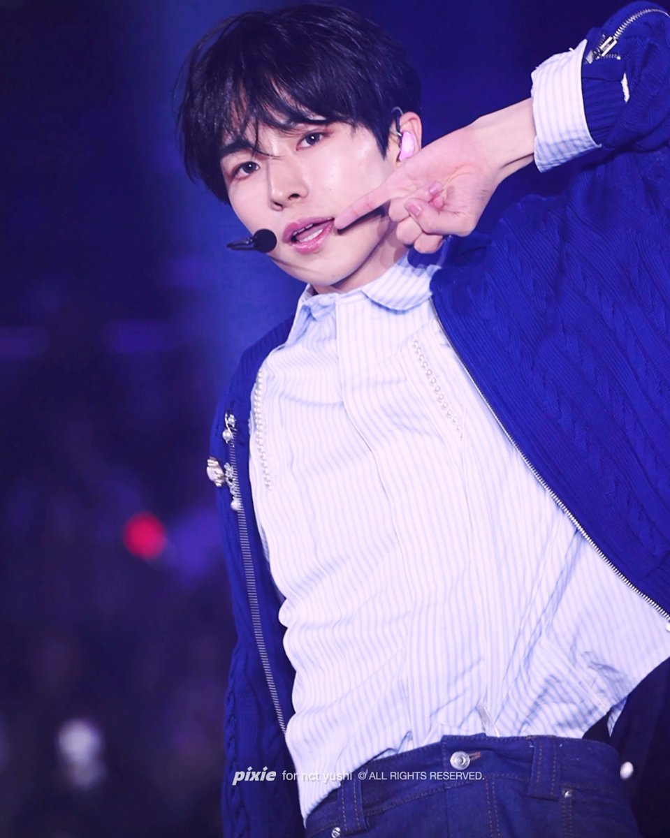 A male performer on stage, wearing a blue jacket over a striped shirt and an earpiece microphone, points to his cheek. The image bears a watermark 'pixie for nct yushi © ALL RIGHTS RESERVED.'