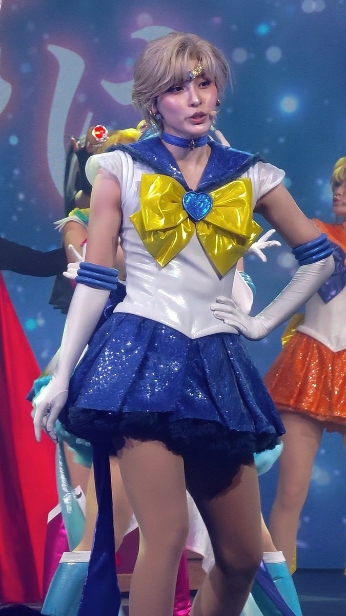 A performer dressed as Sailor Uranus in a blue and white sailor costume with a yellow bow and blue glittery skirt stands on stage.