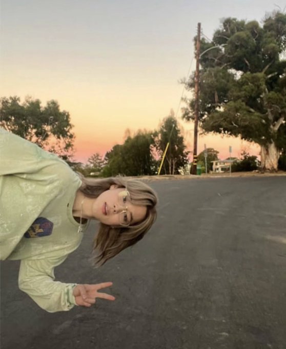 A young woman with glasses and a green sweater taking a peace sign selfie on a street at sunset, with the photo oriented sideways.