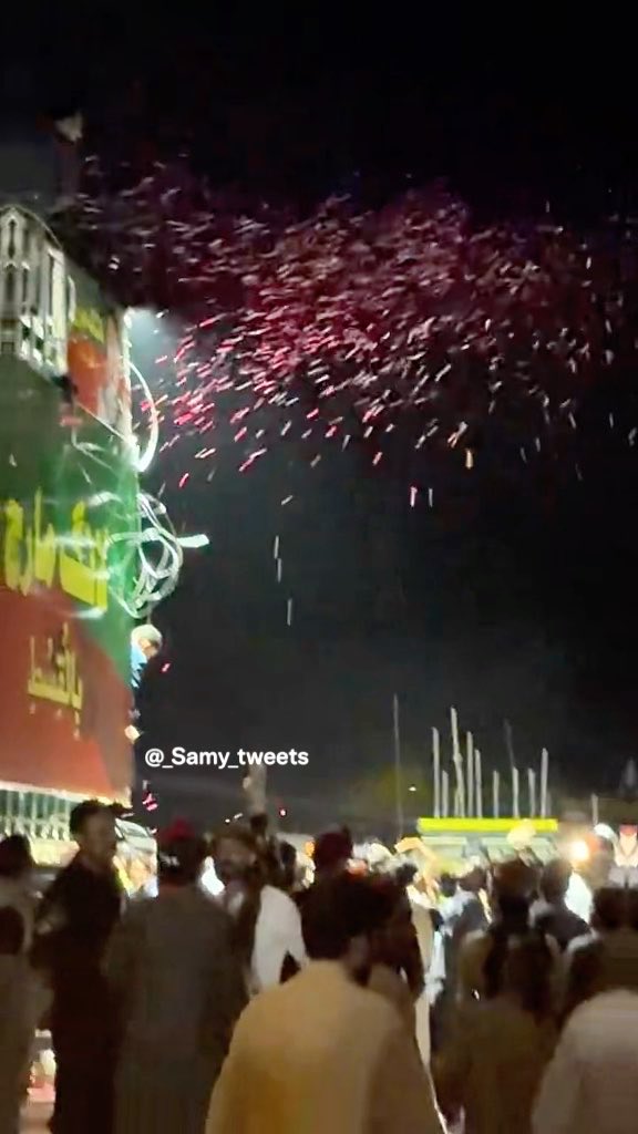 A nighttime outdoor scene showing a crowd of people looking up at fireworks or an aerial display near a building with green and red illuminated signage, with the watermark '@_Samy_tweets' visible.