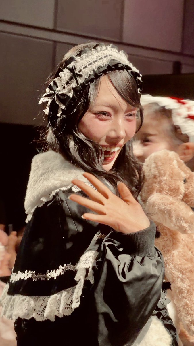 A model named Miu-chan with black-and-white curly hair and dramatic makeup waves during a stage finale, wearing a black Lolita-style outfit with white lace trim and a frilled headband, while holding a teddy bear. The caption relates to the fan service moment captured at Candy Stripper's 30th Anniversary 1st Fashion Show held on October 4, 2026.