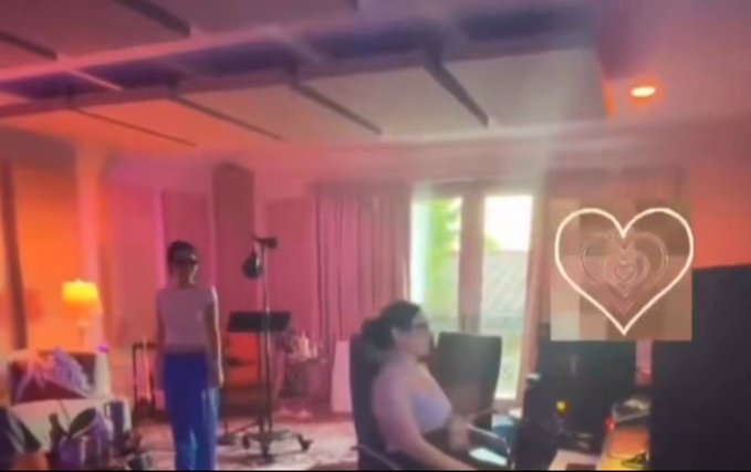 Two people are in a brightly lit room with pink and orange lighting; a heart emoji sticker is overlaid on the right side of the screen.