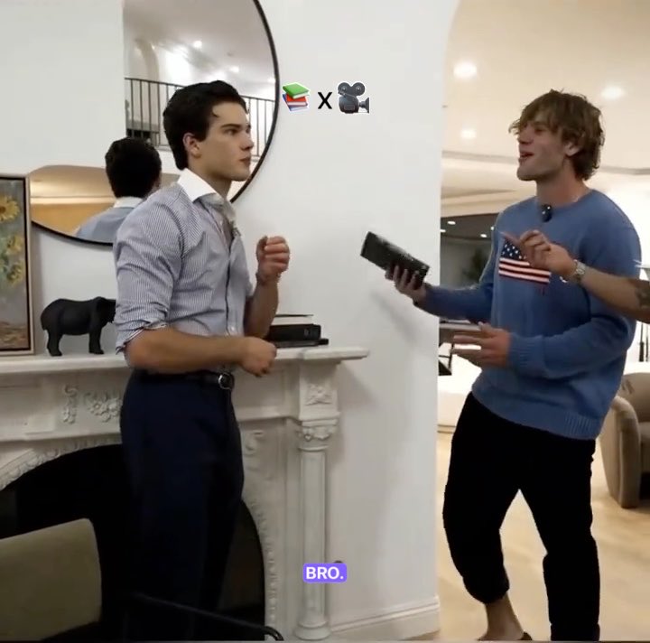 A screenshot from a video showing two men in a modern interior, one in a light blue shirt and the other in a blue sweater with an American flag patch, with a humorous emoji overlay (books and a camera) and the caption 'BRO.'