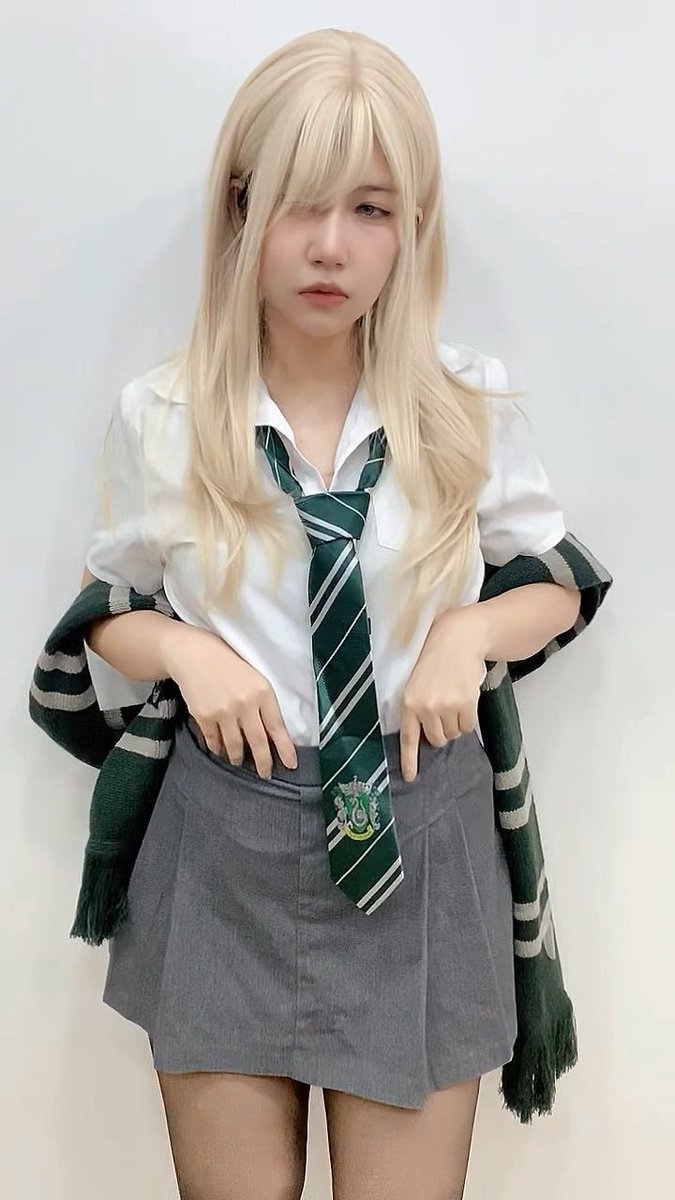 A person with long blonde hair wearing a Slytherin-style cosplay outfit featuring a white shirt, green and silver striped tie, and grey skirt, posed against a plain wall.