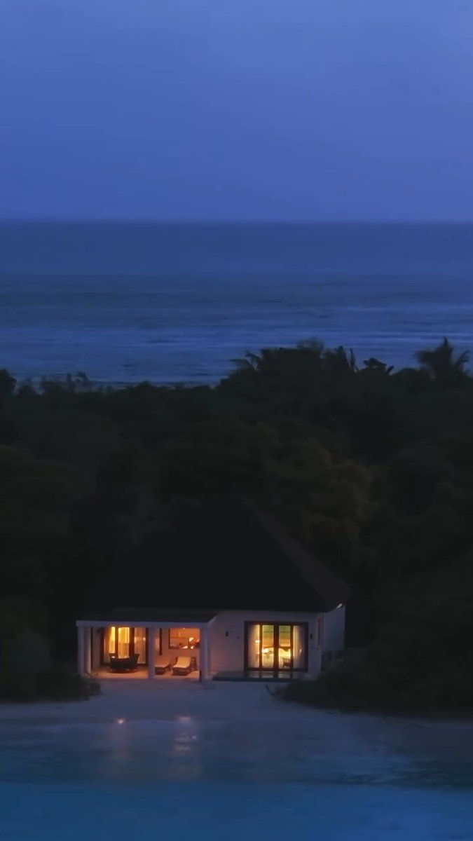 A solitary, illuminated modern cabin with a thatched roof sits by an infinity pool overlooking the dark ocean at twilight.