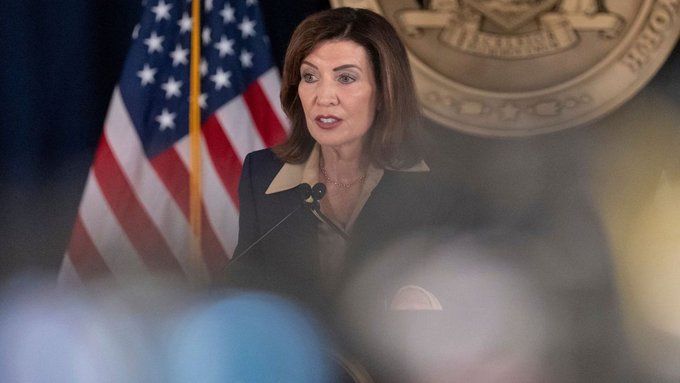 A woman with shoulder-length brown hair speaking at a podium with microphones, with an American flag and an official seal visible behind her.