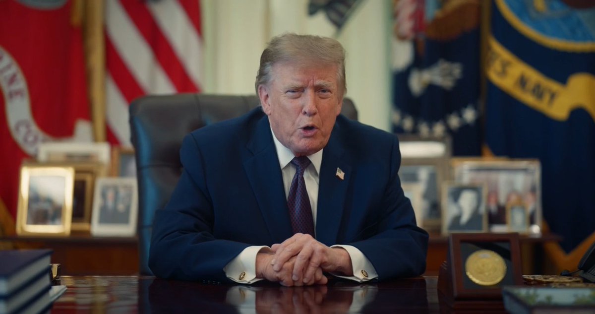 Former US President Donald Trump sits at the Resolute desk in the White House, wearing a dark blue suit and tie, with American flags and military banners visible behind him.
