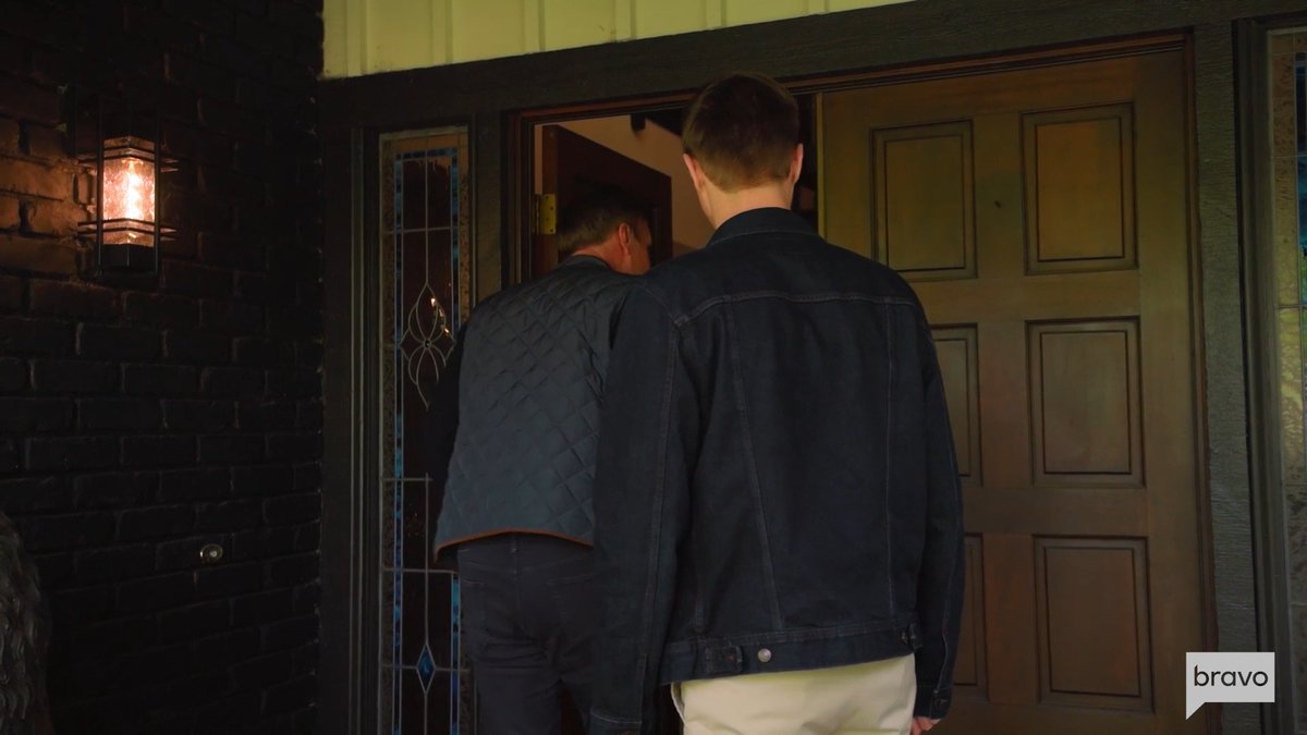 Two men, viewed from behind, approach an open doorway of a house with a dark wooden door and stained glass side panel, illuminated by a glowing outdoor lantern mounted on a brick wall. The Bravo network logo in the corner reinforces this is a scene from "Flipping Out," fitting the post's tease that even veteran Jeff Lewis is in for surprises in a new episode.