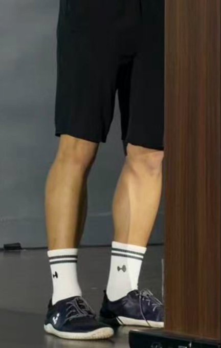 A person stands wearing black shorts, white Under Armour crew socks, and navy sneakers, showing off muscular calves.