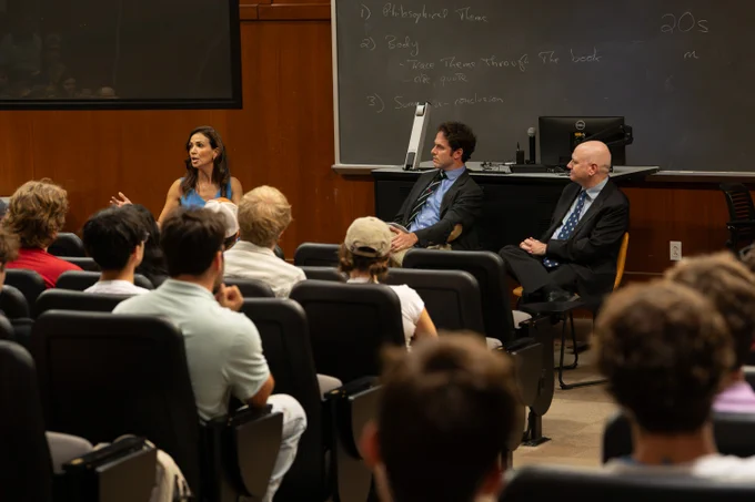 An eye-level shot shows a panel discussion in a university lecture hall. A woman with long brown hair, wearing a bright blue sleeveless top, sits on the left side of the stage, gesturing with her hands as she speaks to an audience seated in tiered rows of black chairs. To her right, two men sit in chairs facing the audience. The man in the middle, wearing a grey blazer, a light blue button-down shirt, a dark tie with red stripes, and khaki pants, looks toward the woman. The man on the far right, wearing a black suit, a patterned blue tie, and dark pants, sits with his hands clasped, looking forward. Behind them is a large chalkboard with white handwritten notes about essay structure, including terms like "Philosophical Theme," "Body," and "Summary-conclusion." A black computer monitor and desk are positioned behind the panel. The audience of students is seen from behind, facing the stage.