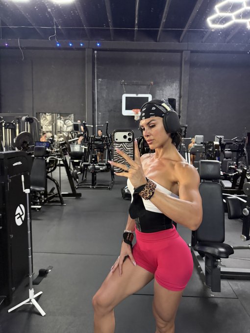 A muscular woman takes a mirror selfie in a gym, wearing a black bandana, leopard-print wristband, black and white top, and pink shorts.