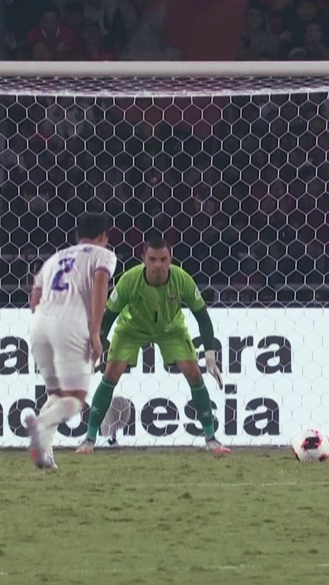 A soccer goalkeeper in a green #1 jersey stands ready in front of the net, facing an opposing player wearing a white #23 jersey with a ball nearby, during what appears to be a #FIFAASEANCup match that Emil Audero is heroically defending for Indonesia.