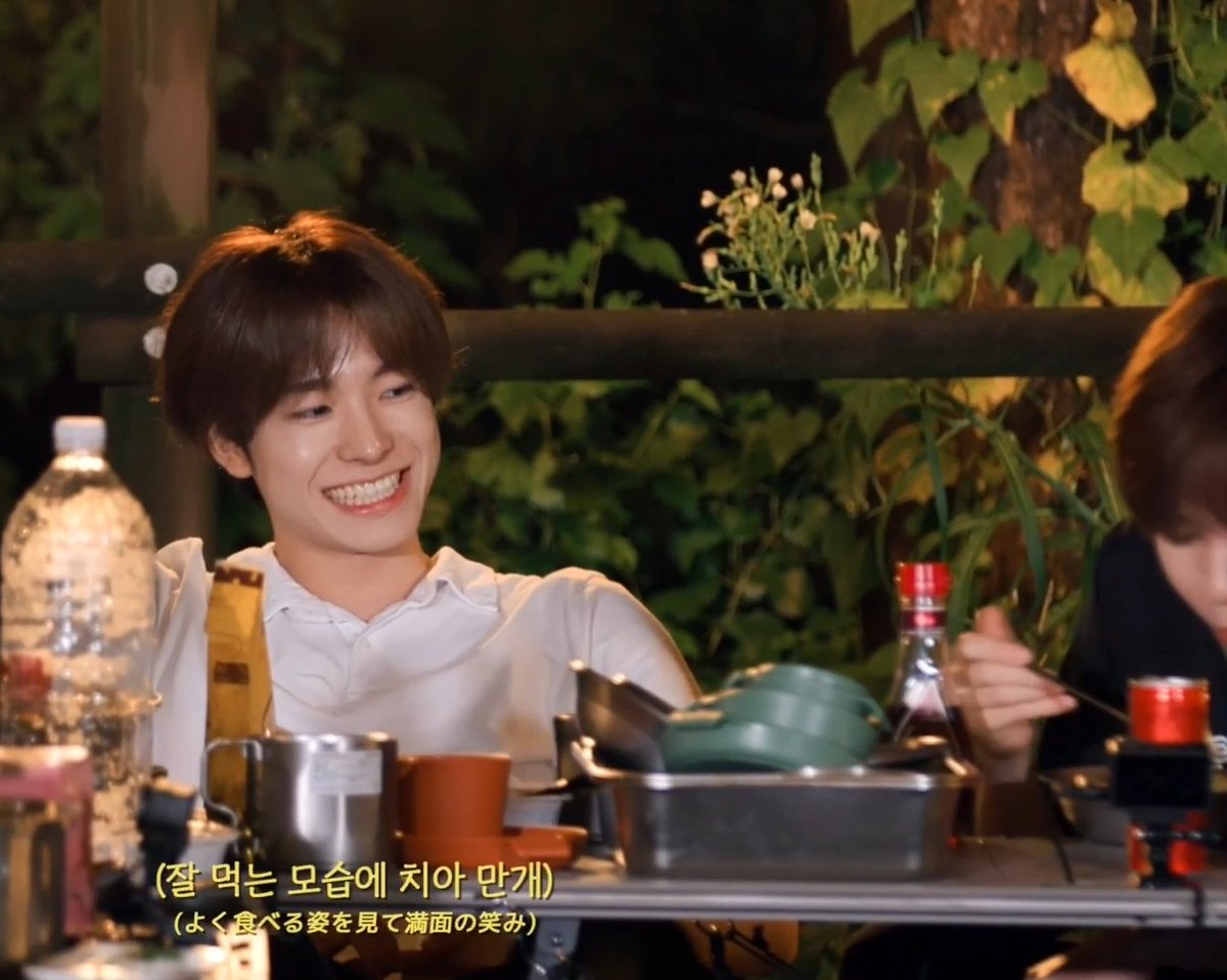 A man with short dark hair smiles broadly while sitting at an outdoor table with cooking equipment; Korean and Japanese subtitles read '(잘 먹는 모습에 치어 만개)' and '(よく食べる姿を見て満面の笑み)'.