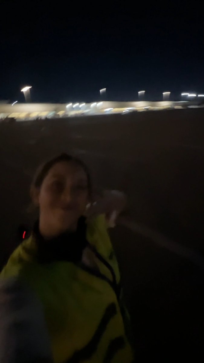 A blurry nighttime selfie shows a smiling woman in a high-visibility yellow jacket, with illuminated stadium lights visible in the background, capturing a behind-the-scenes moment as she joins a team preparing for a special delivery to Atlanta.