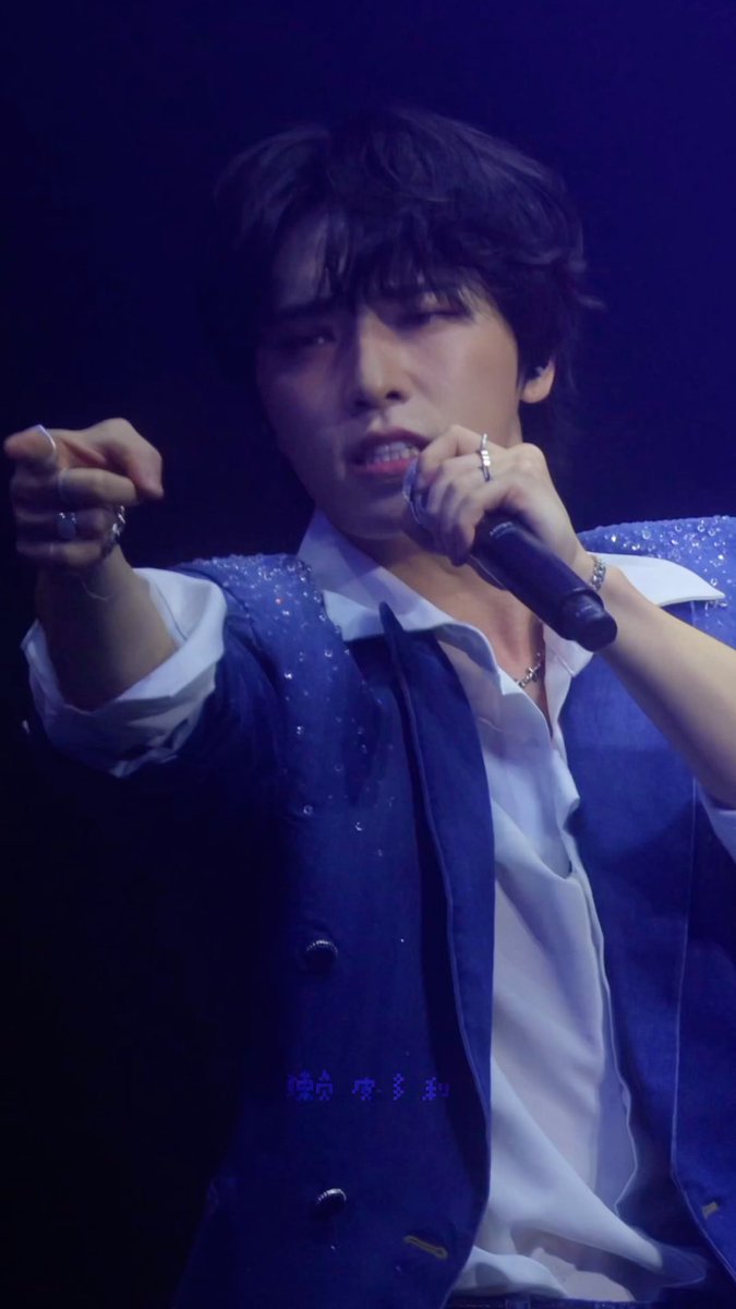 A male K-pop performer on stage singing into a microphone while wearing a sparkly blue vest over a white shirt, accessorized with rings and bracelets, with Chinese text overlay at the bottom.