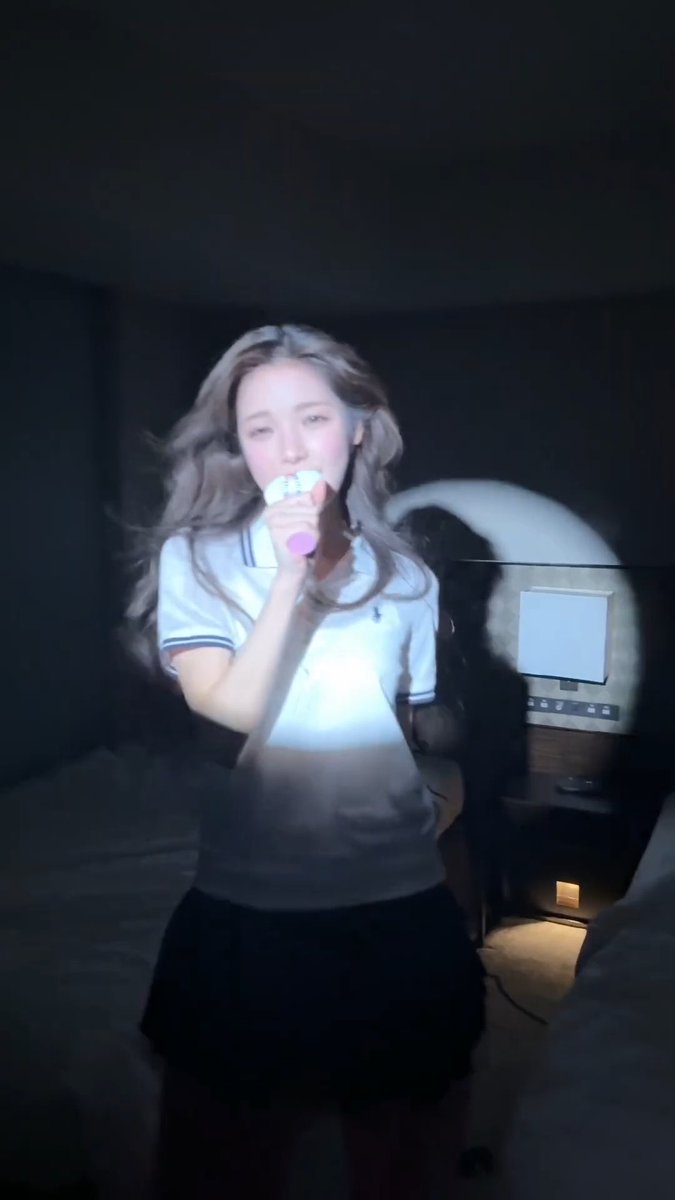 A young woman singing into a pink microphone in a dimly lit room, wearing a white polo shirt and black skirt, with her image partially blurred.