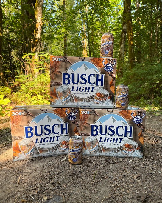 Three cases of Busch Light (each labeled 30 cans) are stacked into a pyramid formation on a dirt forest path, with individual Busch Light cans placed on top and in front of the display—fittingly dubbed the "8th wonder of the world" given the beer brand's cult following among outdoor enthusiasts.
