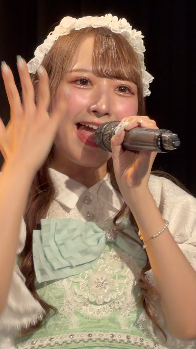 A young performer in a white frilled headband and mint green lace dress sings into a microphone while waving to the audience, her radiant smile matching the affectionate message "ななえは特別です #ななへをみんなへ" ("Nanae is special #NanaetoEveryone") shared in the caption.